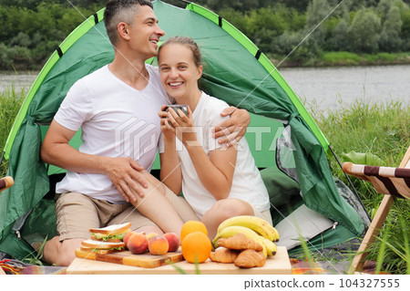 Happy loving smiling family resting outdoor. Romantic woman and man wearing casual clothing sitting at tent near the river drinking coffee or tea, hugging, enjoying time together. 104327555