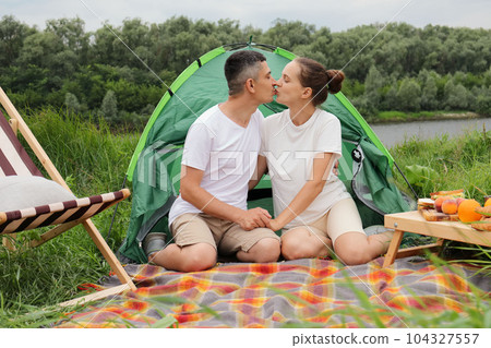 Loving man and woman sitting near tent at lake or river, having picnic in open air, couple hugging and kissing, being on romantic weekend on nature. Loving man and woman sitting near tent at lake or river, having picnic in open air, couple hugging and kissing, being on romantic weekend on nature. 104327557
