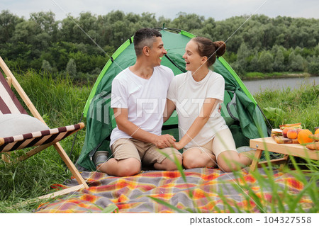 Loving romantic couple woman and man wearing casual clothing sitting at tent near the river holding hands looking at each other enjoying beautiful nature and time spent together. Loving romantic couple woman and man wearing casual clothing sitting at tent near the river holding hands looking at each other enjoying beautiful nature and time spent together. 104327558