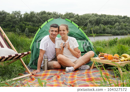 Happy family at weekend, wife and husband relaxing near the river, having picnic, enjoying coffee or tea, looking at camera with smile. Happy family at weekend, wife and husband relaxing near the river, having picnic, enjoying coffee or tea, looking at camera with smile. 104327572