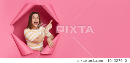 Excited positive brunette woman wearing striped top looking through ripped pink paper hole pointing at advertisement area saying wow empty space for promotion. Excited positive brunette woman wearing striped top looking through ripped pink paper hole pointing at advertisement area saying wow empty space for promotion. 104327640