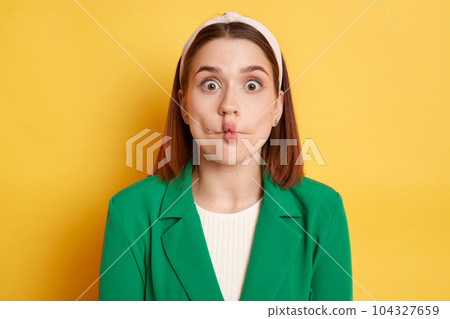 Funny optimistic woman wearing green jacket making fish lips grimacing having fun looking at camera with big eyes posing isolated over yellow background. Funny optimistic woman wearing green jacket making fish lips grimacing having fun looking at camera with big eyes posing isolated over yellow background. 104327659