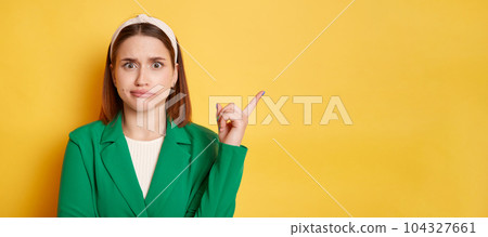 Confused puzzled woman wearing green jacket posing isolated over yellow background pointing away at copy space, advertisement area mockup. Confused puzzled woman wearing green jacket posing isolated over yellow background pointing away at copy space, advertisement area mockup. 104327661
