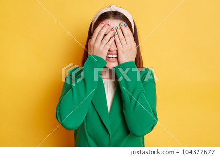 Smiling woman wearing green jacket posing isolated over yellow background hiding her eyes covering face with palms positive emotions. Smiling woman wearing green jacket posing isolated over yellow background hiding her eyes covering face with palms positive emotions. 104327677