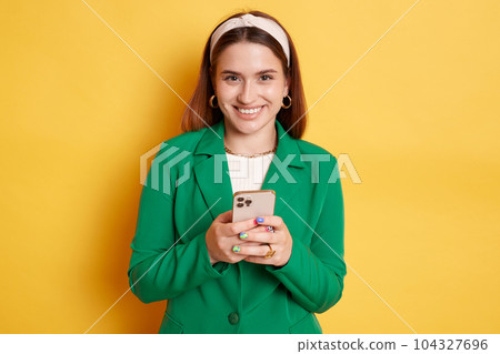 Cute attractive happy woman wearing green jacket posing isolated over yellow background holding mobile in hands checking social networks. Cute attractive happy woman wearing green jacket posing isolated over yellow background holding mobile in hands checking social networks. 104327696