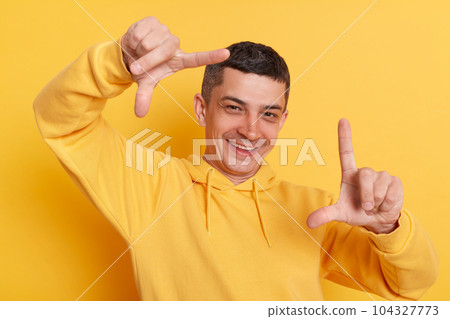 Cheerful photographer. Smiling handsome man wearing casual hoodie looking at camera with satisfied expression trough frame of fingers posing isolated over yellow background. 104327773