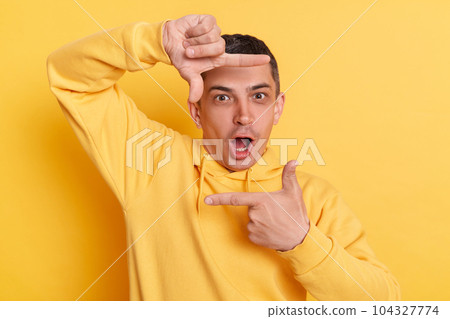 Shocked photographer. Surprised astonished man wearing casual hoodie making frame of fingers looking at camera with open mouth posing isolated over yellow background. 104327774