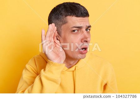Confused man wearing casual hoodie standing with hands near ear trying to listen silent talking posing isolated over yellow background. Confused man wearing casual hoodie standing with hands near ear trying to listen silent talking posing isolated over yellow background. 104327786