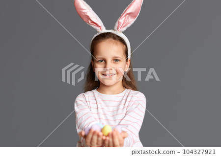 Photo of satisfied little girl wearing bunny ears holding Easter eggs, found many testicles during Easter hunt, rejoicing, showing her result, isolated on gray background 104327921