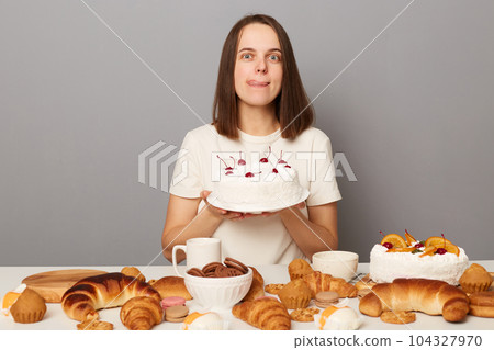 Portrait of hungry sugary addicted woman with brown hair wearing white T-shirt sitting at table isolated over gray background, holding cake, showing tongue out. 104327970