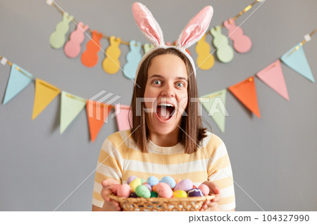 Indoor shot of amazed excited woman wearing rabbit ears holding colorful Easter eggs in wicker basket isolated on gray decorated background, screaming with happiness, waiting Easter celebration Indoor shot of amazed excited woman wearing rabbit ears holding colorful Easter eggs in wicker basket isolated on gray decorated background, screaming with happiness, waiting Easter celebration 104327990