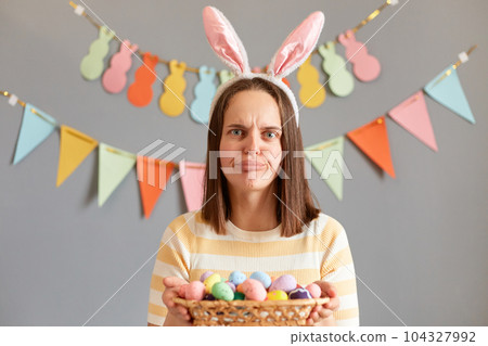 Indoor shot of disappointed sad upset woman wearing rabbit ears holding colorful Easter eggs in basket, looking at camera with sorrow, celebrating holiday alone, isolated on gray decorated background Indoor shot of disappointed sad upset woman wearing rabbit ears holding colorful Easter eggs in basket, looking at camera with sorrow, celebrating holiday alone, isolated on gray decorated background 104327992