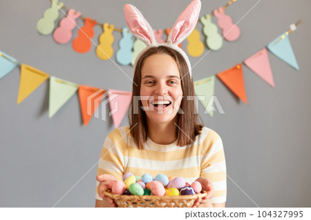 Portrait of attractive happy brown haired woman wearing rabbit ears holding Easter eggs, looking at camera with happy face, painting eggs for holiday, isolated on gray decorated background Portrait of attractive happy brown haired woman wearing rabbit ears holding Easter eggs, looking at camera with happy face, painting eggs for holiday, isolated on gray decorated background 104327995
