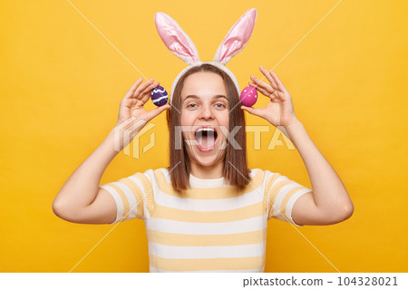 Photo of excited joyful cheerful woman wearing rabbit ears holding colorful Easter eggs, having fun while celebrating, yelling with happy face, isolated on yellow background Photo of excited joyful cheerful woman wearing rabbit ears holding colorful Easter eggs, having fun while celebrating, yelling with happy face, isolated on yellow background 104328021