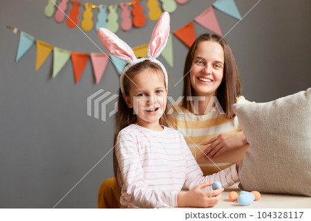 Easter traditions. Photo of little girl wearing rabbit ears with her mommy find eggs under pillow, looking at camera, Easter hunt family posing in home interior with decorations. Easter traditions. Photo of little girl wearing rabbit ears with her mommy find eggs under pillow, looking at camera, Easter hunt family posing in home interior with decorations. 104328117