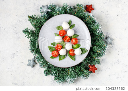Caprese salad in the form of a Christmas tree. Festive tomato mozzarella and basil appetizer on light plate. 104328362