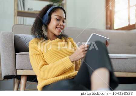 Portrait of an African American with headphones and tablet smiling happily listen music while relaxing on the sofa at home. Portrait of an African American with headphones and tablet smiling happily listen music while relaxing on the sofa at home. 104328527