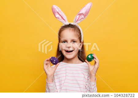 Portrait of excited charming girl with rabbit ears on her head with colored eggs in hands, being in good mood, celebrating spring holiday, posing isolated on yellow background Portrait of excited charming girl with rabbit ears on her head with colored eggs in hands, being in good mood, celebrating spring holiday, posing isolated on yellow background 104328722