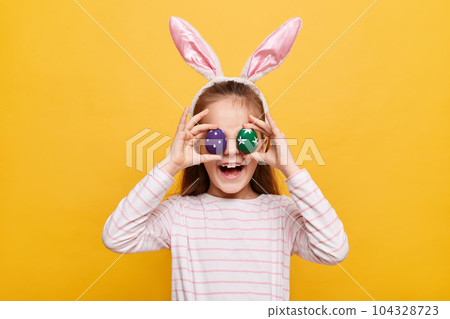 Portrait of funny little girl with rabbit ears with covering her eyes colored purple and green eggs having fun smiling happily posing isolated on yellow background 104328723