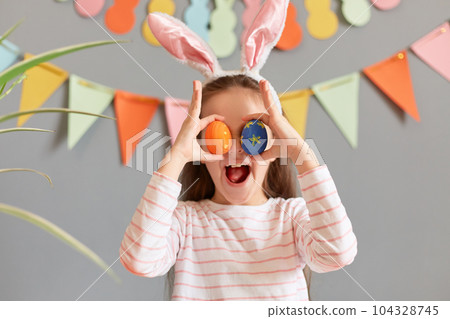 Indoor shot of amazed cute excited little girl wearing rabbit ears, hiding her eyes with Easter eggs, having fun, preparing for holiday, standing against decorated gray wall 104328745