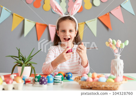 Photo of excited adorable little girl painting aster eggs with brush, wearing bunny ears looking at camera with happiness, celebrating Easter, posing against decorated gray background. 104328760