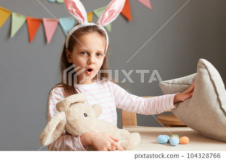 Portrait of shocked surprised little girl wearing rabbit ears being amazed to find so many eggs under pillow on Easter hunt posing in home interior with decorations, looking at camera with big eyes. Portrait of shocked surprised little girl wearing rabbit ears being amazed to find so many eggs under pillow on Easter hunt posing in home interior with decorations, looking at camera with big eyes. 104328766