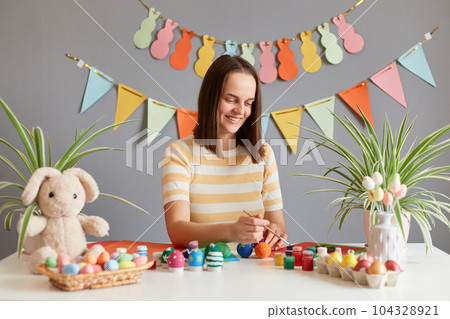 Indoor shot of joyful positive attractive woman with brown hair wearing casual clothing preparing for Easter and coloring eggs, enjoying process, posing against decorated festive wall Indoor shot of joyful positive attractive woman with brown hair wearing casual clothing preparing for Easter and coloring eggs, enjoying process, posing against decorated festive wall 104328921