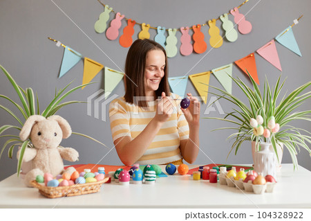 Portrait of smiling cheerful woman with dark straight hair wearing striped shirt painting Easter eggs preparing for holiday, being in good festive mood, posing against decorated festive wall Portrait of smiling cheerful woman with dark straight hair wearing striped shirt painting Easter eggs preparing for holiday, being in good festive mood, posing against decorated festive wall 104328922