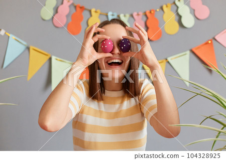 Portrait of amazed laughing excited charming woman with brown hair in casual clothing covering her eyes with Easter eggs, having fun during holiday preparation, posing against decorated festive wall Portrait of amazed laughing excited charming woman with brown hair in casual clothing covering her eyes with Easter eggs, having fun during holiday preparation, posing against decorated festive wall 104328925