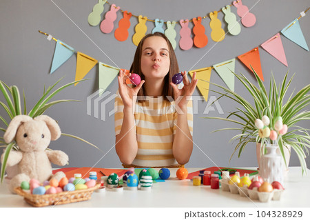 Image of pretty adorable woman with brown hair wearing striped shirt celebrating Easter, holding colored eggs, sending air kissing, congratulating with holiday against decorated festive wall 104328929