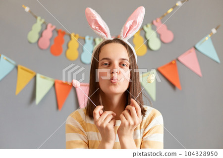 Portrait of romantic pretty woman wearing rabbit ears holding colorful Easter cake pops isolated on gray decorated background, sending air kissing, congratulating with holiday Portrait of romantic pretty woman wearing rabbit ears holding colorful Easter cake pops isolated on gray decorated background, sending air kissing, congratulating with holiday 104328950