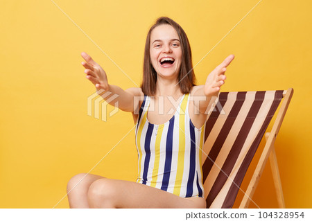 Portrait of friendly happy young woman wearing striped one-piece swimsuit sitting on wooden chair isolated on yellow background, inviting you to resort, saying come into my arms 104328954