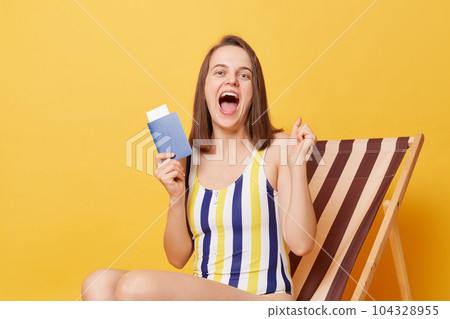 Horizontal shot of amazed excited overjoyed young woman wearing colorful swimsuit, holding tickets and passport in hands, yelling with happiness, sitting on wooden chair isolated on yellow background 104328955