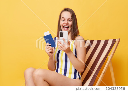 Image of amazed happy young woman wearing striped one-piece swimsuit, holding passport and using cell phone, booking tickets for traveling, sitting on wooden chair isolated on yellow background 104328956