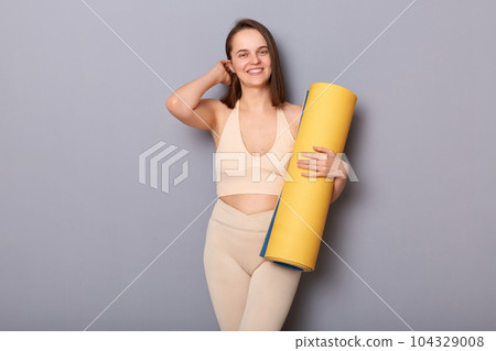 Image of pretty cute charming woman wearing beige top and leggins holding fitness mat isolated over gray background, touching her hair, smiling at camera, visiting gym for improving body shape 104329008