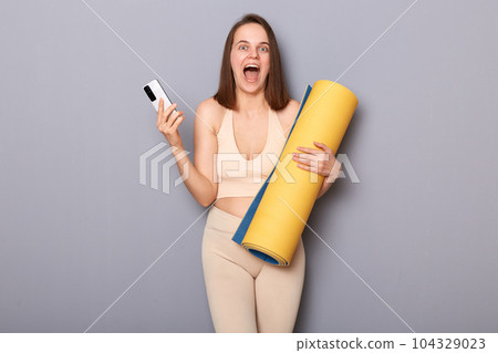 Portrait of excited amazed athletic woman holding rolled up fitness mat and mobile phone, screaming with happiness, winning free fitness gym membership. 104329023