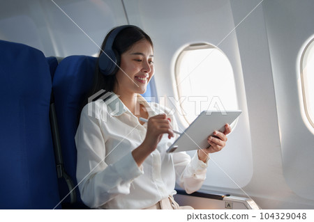 Young Asian attractive business woman passenger sitting on business class luxury plane while wearing wireless headphone for relax and working with tablet during the flight. 104329048