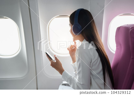 Young Asian attractive business woman passenger sitting on business class luxury plane while wearing wireless headphone and using smart phone mobile for relax during the flight. Young Asian attractive business woman passenger sitting on business class luxury plane while wearing wireless headphone and using smart phone mobile for relax during the flight. 104329312