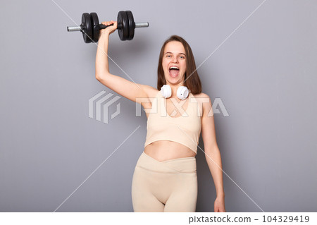 Portrait of crazy excited strong woman wearing top and leggins holding barbell in hand raised her arm doing exercise for her triceps and biceps isolated on gray background Portrait of crazy excited strong woman wearing top and leggins holding barbell in hand raised her arm doing exercise for her triceps and biceps isolated on gray background 104329419