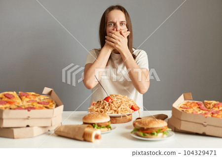 Portrait of sick woman with brown hair wearing white T-shirt sitting at table isolated over gray background, feeling nausea after overeating junk food. 104329471