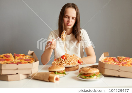 Indoor shot of frustrated sad woman with brown hair wearing white T-shirt sitting at table isolated over gray background, eating pasta, being disappointed, being not hungry. 104329477