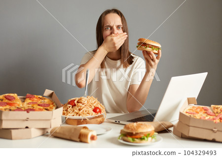 Portrait of sick overeating woman sitting at table in front of notebook isolated over gray background, holding sandwich, feels bad and nausea, covering mouth with palm. 104329493