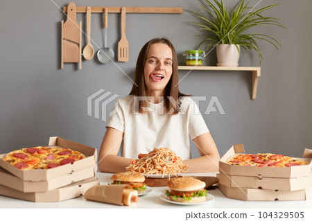 Indoor shot of cheerful attractive woman with brown hair wearing white casual T-shirt sitting at table in kitchen, feels hungry, looking at camera, showing tongue our, want to eat fast food. Indoor shot of cheerful attractive woman with brown hair wearing white casual T-shirt sitting at table in kitchen, feels hungry, looking at camera, showing tongue our, want to eat fast food. 104329505
