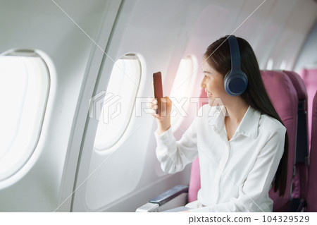 Young Asian attractive business woman passenger sitting on business class luxury plane while wearing wireless headphone and using smart phone mobile for relax during the flight. 104329529