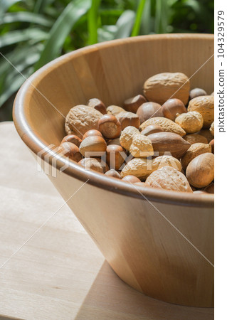 Assorted various nuts in a big wooden bowl against a leafy green background 104329579