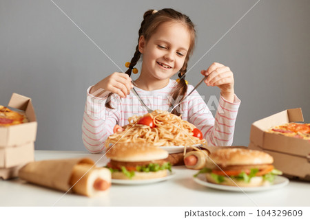 Portrait of satisfied little girl with pigtails sitting at table with big variety of junk food, eating pasta with satisfied expression, isolated over gray background 104329609