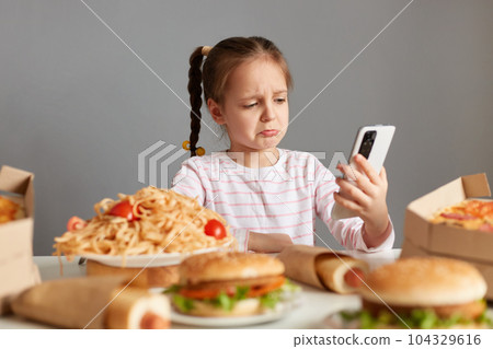 Indoor shot of sad crying little girl with pigtails sitting at table pizza, hot dogs, burgers, talking via video call, keeps diet, can't eat delicious fast food, isolated over gray background Indoor shot of sad crying little girl with pigtails sitting at table pizza, hot dogs, burgers, talking via video call, keeps diet, can't eat delicious fast food, isolated over gray background 104329616
