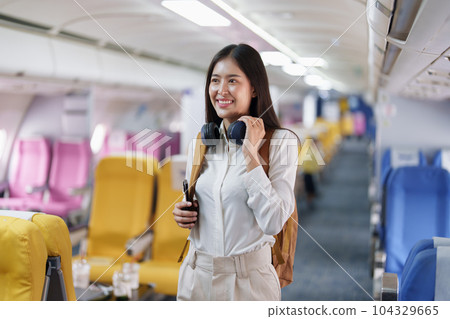 Young asian attractive woman travel by airplane, Passenger wearing headphone putting hand baggage in lockers above seats of plane. Young asian attractive woman travel by airplane, Passenger wearing headphone putting hand baggage in lockers above seats of plane. 104329665