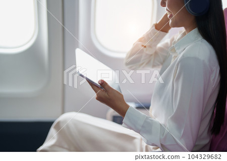 Young Asian attractive business woman passenger sitting on business class luxury plane while wearing wireless headphone for relax and working with tablet during the flight. Young Asian attractive business woman passenger sitting on business class luxury plane while wearing wireless headphone for relax and working with tablet during the flight. 104329682