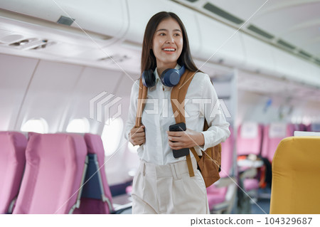 Young asian attractive woman travel by airplane, Passenger wearing headphone putting hand baggage in lockers above seats of plane. Young asian attractive woman travel by airplane, Passenger wearing headphone putting hand baggage in lockers above seats of plane. 104329687